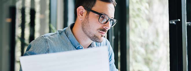 Man with light skin, dark hair and glasses holds paper and looks off screen