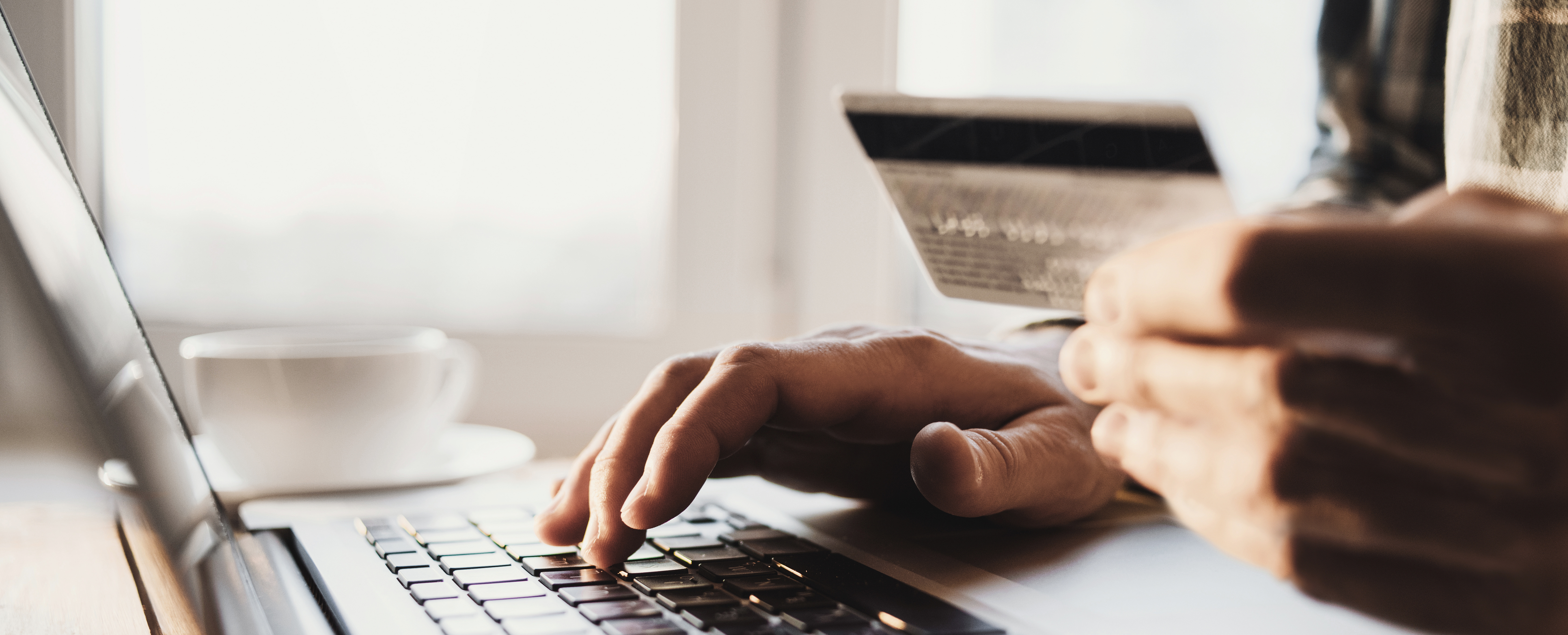 A person holds his credit card in his hands while typing on a laptop on the counter with a coffee cup.