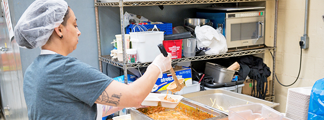 Woman with hairnet and safety gloves helps serve food on tray for Meals on Wheels of Lancaster