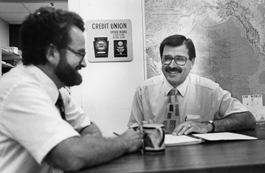 Two founders of Mennonite Mutual Aid with bowties sitting down at table with coffee mugs talking and smiling 