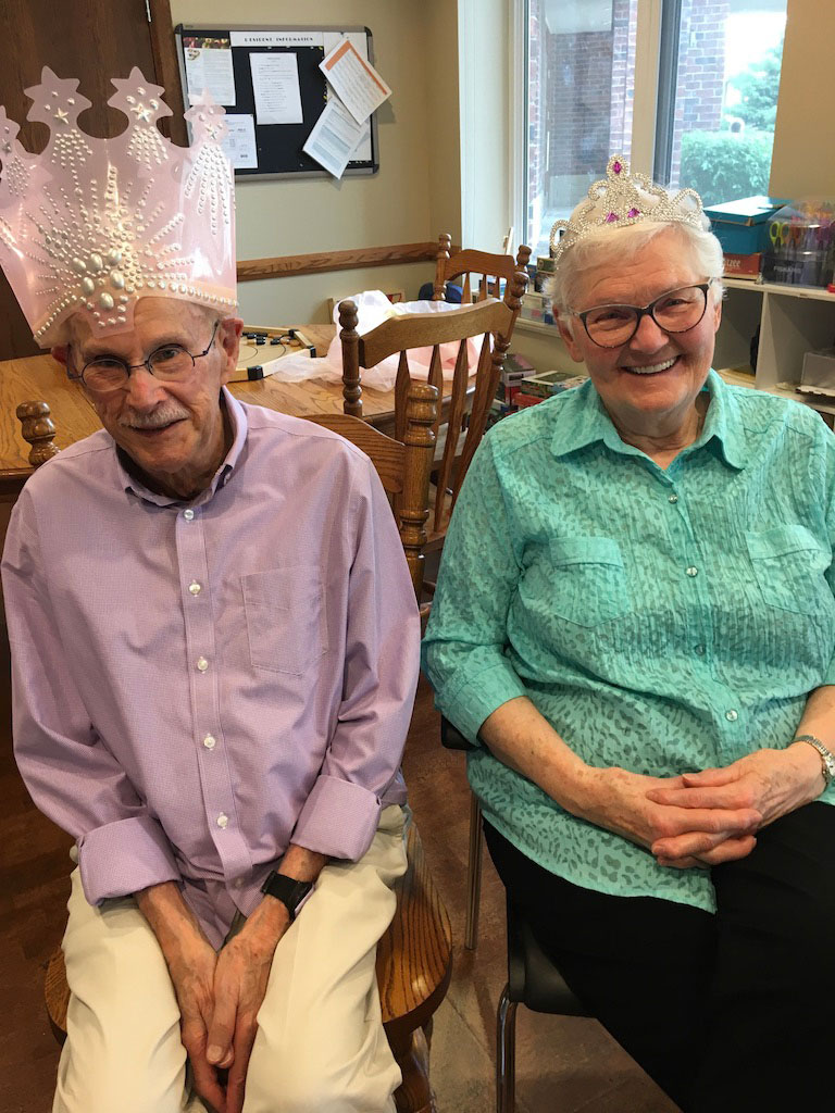 An elderly couple of a man and a woman sit, posing for a photo. They are both wearing silly hats for a party