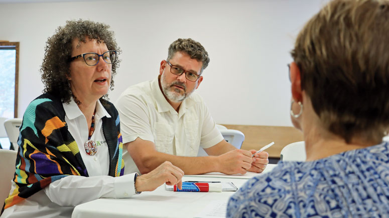 Three people in a conversation. A woman in the foreground, her back to the camera, listens while Marlene Kroeker in the background speaks, as a man next to her looks on