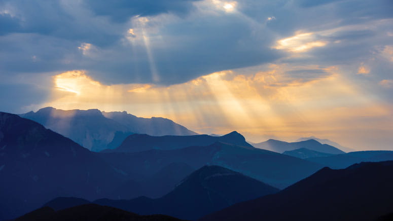 A landscape photo of a mountain range. Rays of sun push through clouds in the horizon