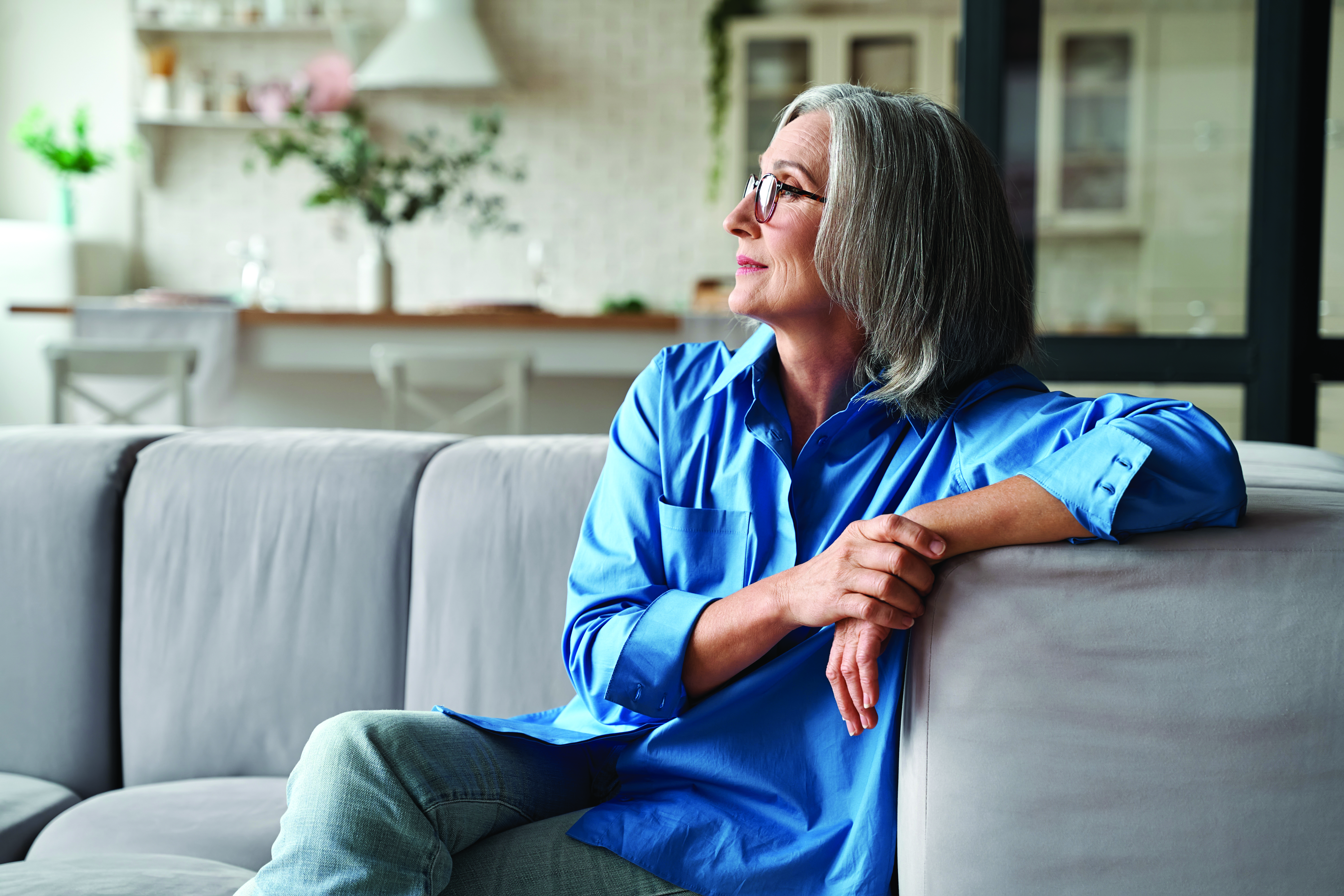 Middle aged white woman with gray hair looks off into the distance while sitting on a couch.