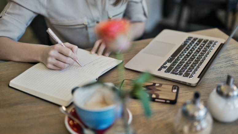 Person writing in a notebook on a desk with a laptop and coffee.