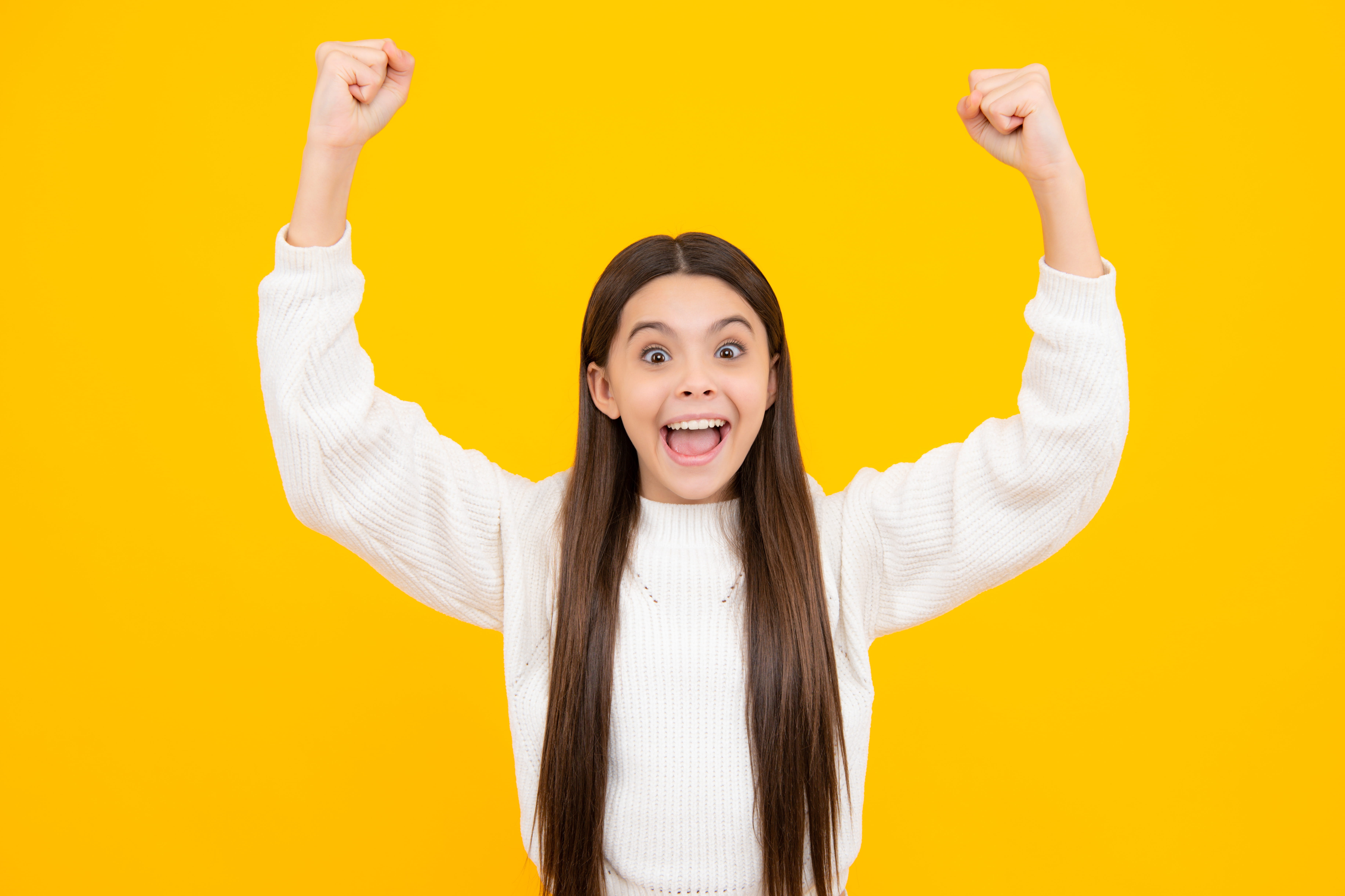 Preteen girl with dark hair raising both fists in the air with enthusiasm and excitement like she won in front of a yellow background.