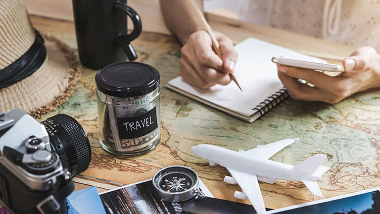 Person surrounded by travel-themed items with jar of money that says "travel"