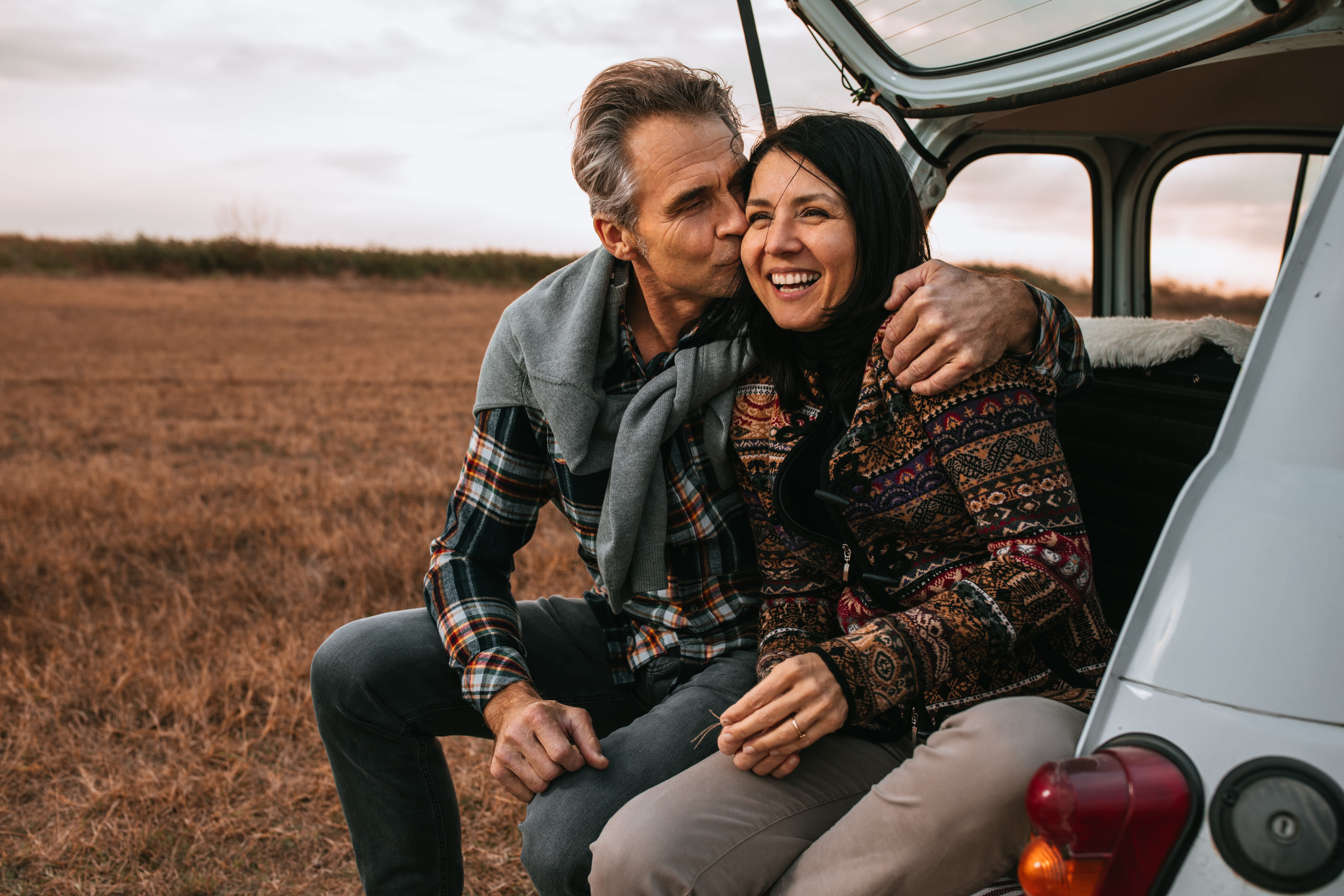 Middle-aged couple with sweaters sit on the back of a car looking at a fall scene.