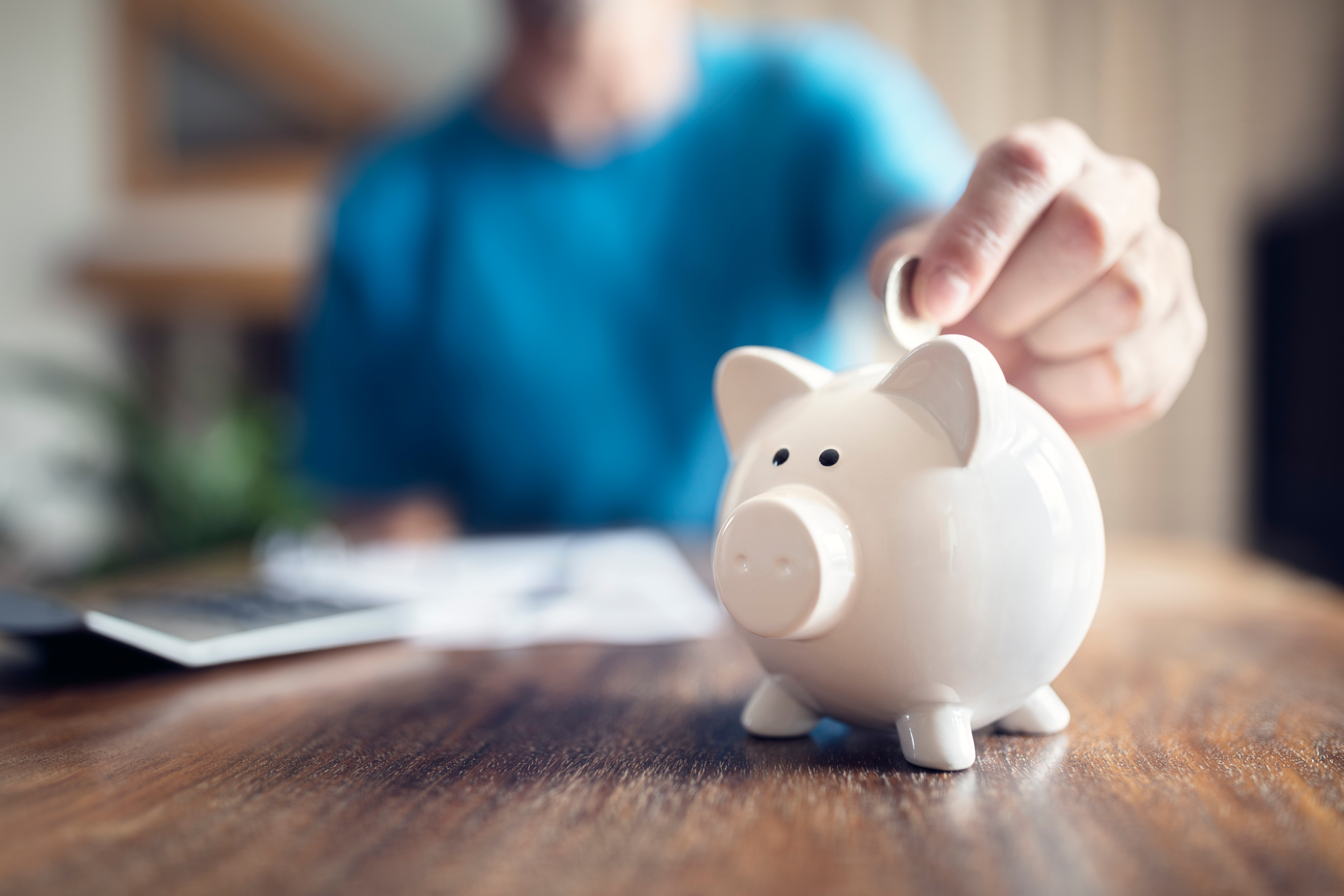 A man in a blue shirt puts a coin in a piggy bank on a wooden table.