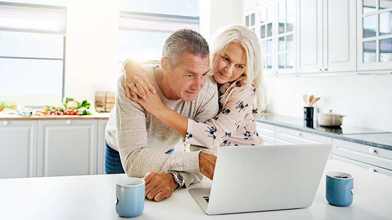 An older couple look at a computer together in a bright white kitchen.