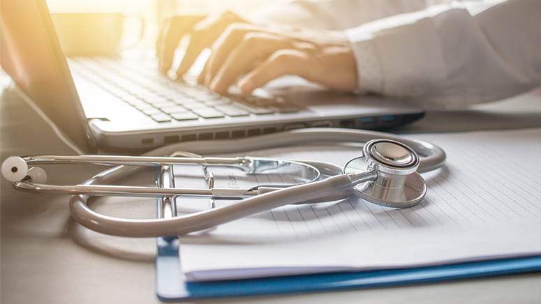 Stethoscope curled on top of notebook with person typing on laptop keyboard in background